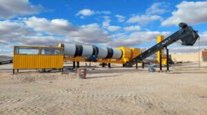 Usine continue d’asphalte de couleur jaune installée sur un site désertique, avec un tambour rotatif et une bande transporteuse inclinée marquée "ALFA", sous un ciel bleu partiellement nuageux.