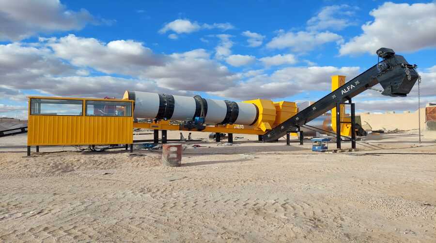 Blog 2 Usine continue d’asphalte de couleur jaune installée sur un site désertique, avec un tambour rotatif et une bande transporteuse inclinée marquée "ALFA", sous un ciel bleu partiellement nuageux.